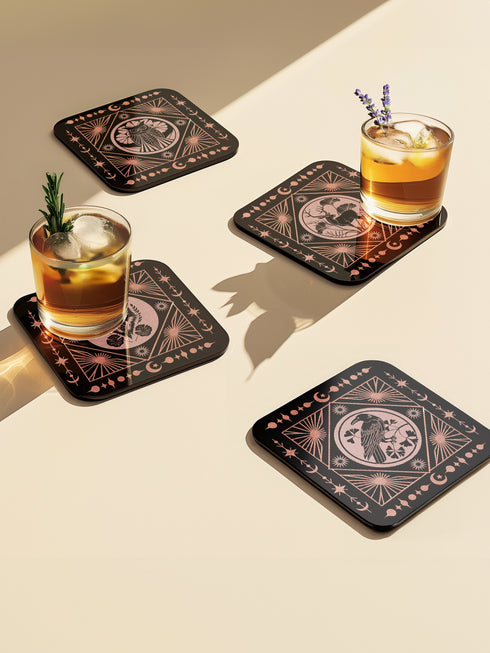 A set of four black, high-gloss square coasters featuring intricate rose-gold metallic illustrations of an owl, raven, stag, and wolf framed by celestial and botanical motifs. Made from durable masonite hardboard with cork backing for slip-resistant protection.